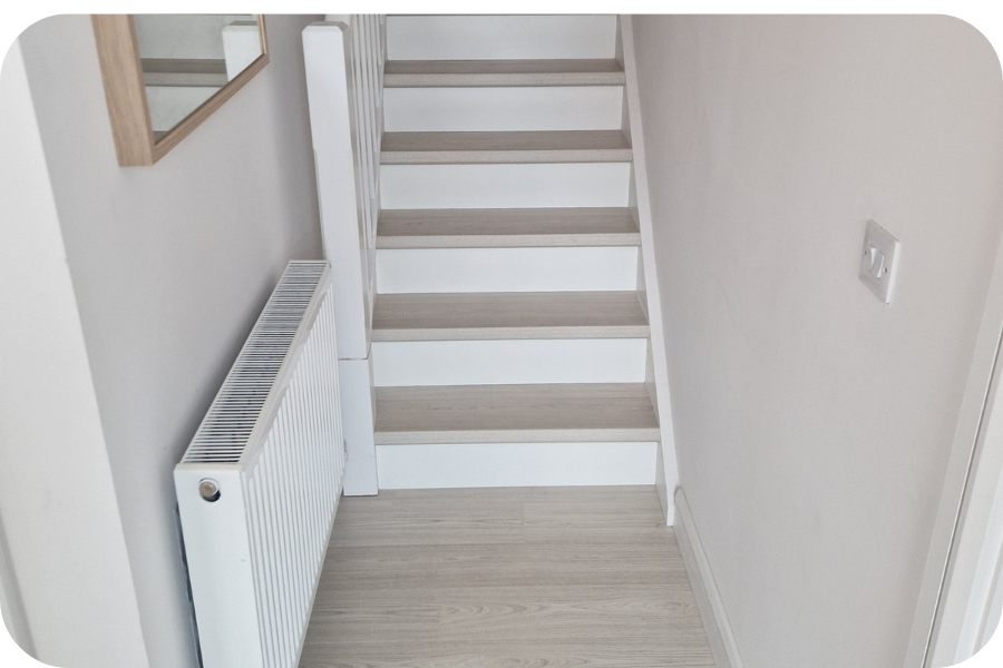 A narrow hallway with light wood flooring, a white radiator on the left wall, a mirror above it, and a staircase with white risers and light wood treads leading upward.