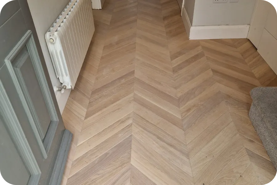 A hallway with light wood chevron-patterned flooring, a white radiator on the left wall, and a gray door partially visible in the foreground. Carpeted stairs are on the right side of the image.