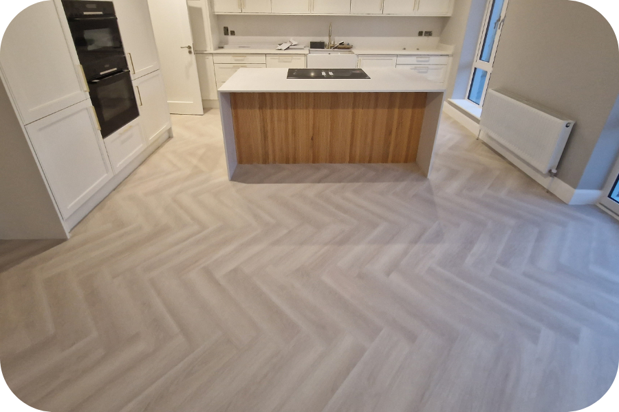 Modern kitchen with white cabinets, an island featuring a wood accent, and light herringbone-patterned flooring. There are built-in ovens, a radiator, and large windows providing natural light.