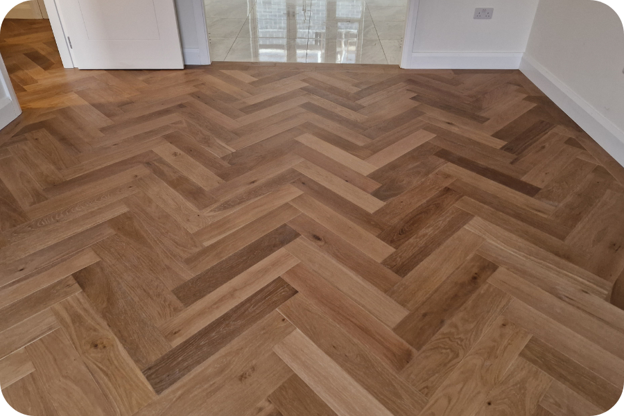A room with light brown herringbone wood flooring, white walls, and an open door leading to a space with glossy beige tiles. The corners of the image are rounded.