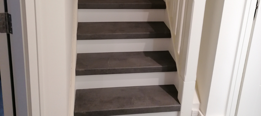 A set of indoor stairs with dark gray steps and white risers, surrounded by white walls and a white banister on the right side. The floor at the base is also gray.