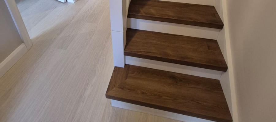 A small staircase with dark wooden steps and white sides meets a light-colored, wood-patterned floor in a modern, clean interior hallway. An open door can be seen in the background.